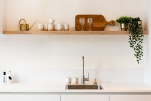 Mat witte keuken greeploos met wandplank voor keukenaccessoires als zand aardetinten kopjes, plantjes en een houten borrelplank Mat witte keuken greeploos met wandplank voor keukenaccessoires als zand aardetinten kopjes, plantjes en een houten borrelplank